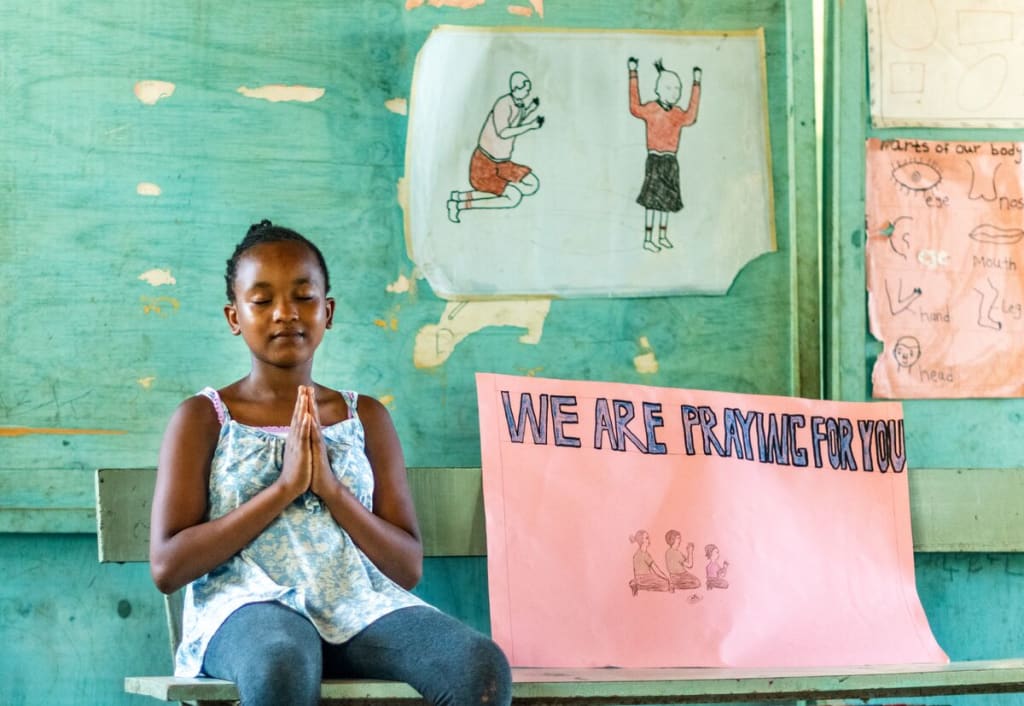 Girl sits on a bench with her eyes closed and hands in prayer position. Beside her is a sign that says, "We are praying for you."